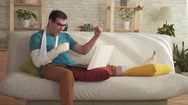 Expressive young man with a broken arm and leg uses a laptop and learns about the win slow mo