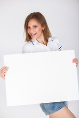 Naklejka premium Beautiful young gir in denim shorts and a T-shirth on a white background holding big white blank card. Pretty student girl having fun and holding white empty signboard background with copy space.
