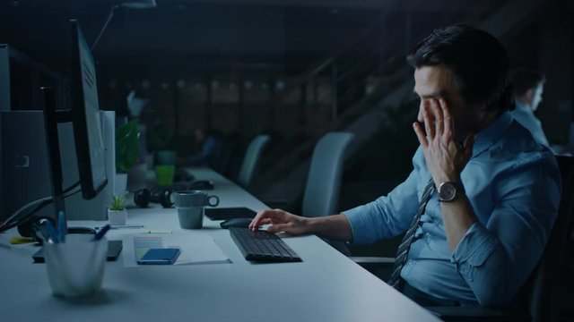 In the Office at Night Overworked Tired Office Worker Uses Desktop Computer Yawning, Drinks Coffee. Tired Exhausted Businessman Finishing Important Project