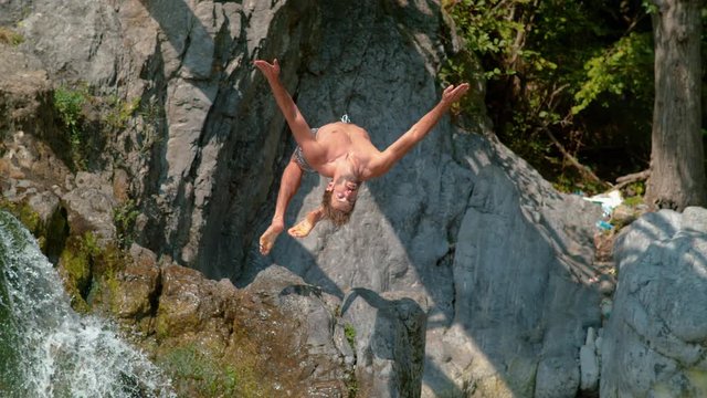 SLOW MOTION: Breathtaking shot of an athletic Caucasian man doing an epic backflip from a towering waterfall. Sporty male tourist diving into the refreshing crystal clear pond on a sunny summer day.