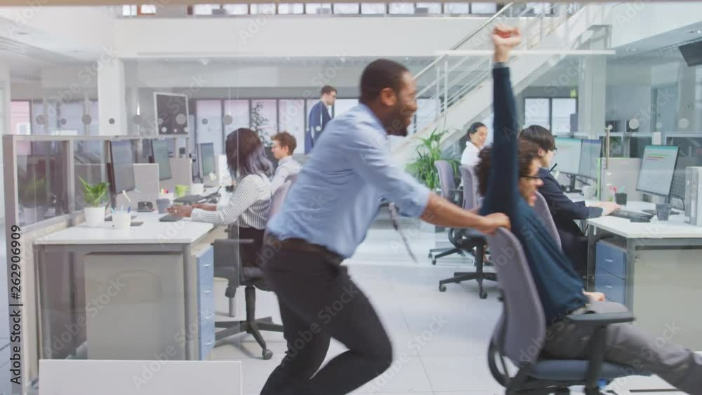 Poster Cheerful Employee Pushes His Handsome Colleague on a Chair Next to Rows of Desks with Diverse Business People Working on Desktop Computers in Modern Office Space. 