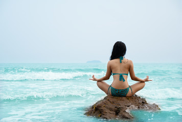 Natural background of Asian female in bikini sitting on rock and having yoga and meditation in sea with blue sky