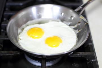Two eggs frying sunny-side up in a stainless steel pan on the stove.