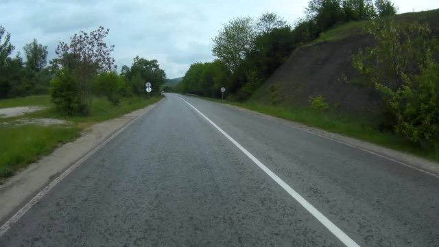 Video background shot in motion from car backwards - 60 fps. An asphalt road outside the city. Green trees along the road. Cloudy, rainy weather.