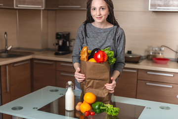 Woman holding full paper bag with products in hands on the background of the kitchen. Healthy and fresh organic food
