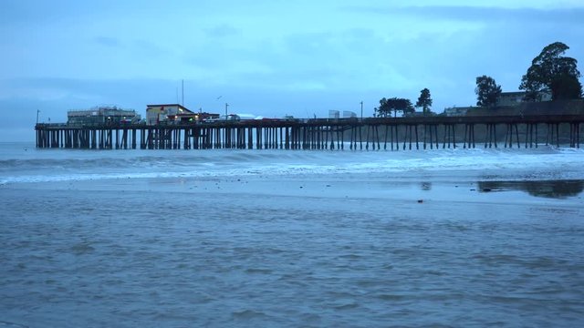Camera zooming in on Capitola Beach Pier in Santa Cruz, California. A stream of water flowing into the ocean in the foreground.