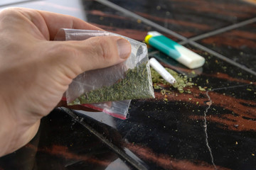 a man's hand holds a sachet with a weed, a lighter and a narcotic cigarette in the background