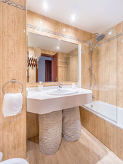 Clean bright bathroom interior with a big mirror,bathtub,white sink and marble tiles. Original designed hotel space with modern pieces