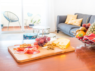 Table with fruit and food in a stylish bright living room with sofa, wooden table, side tables, tv,plants and big window to a terrace with mountain view. Healthy lifestyle concept.