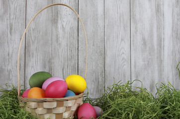 Painted eggs in basket with dry green grass against white wooden wall. Easter background or egg hunt concept. Pink, yellow, green, orange eggs, hay, side view