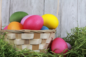 Painted eggs in basket with dry green grass against white wooden wall. Easter background or egg hunt concept. Pink, yellow, green, orange eggs, hay, side view, close-up