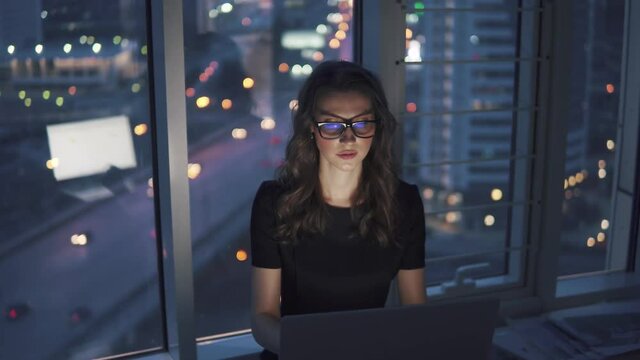 girl works in office in late evening against background of lights of the night of the city. young business woman with a monitor reflection in glasses
