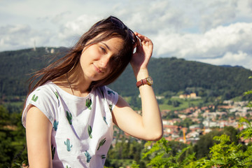 A beautiful smiling attractive girl sits in the light of the sun holding sunglasses against a background of green leaves, mountains and forest in the Bergamo, at the foothills of the Alps, Italy.