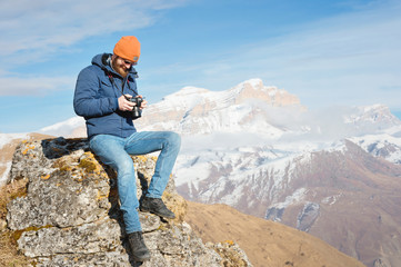 Portrait bearded male photographer in sunglasses and warm jacket with reflex camera in his hands looks at the photos on the camera while sitting on stone in the background of snow-capped mountains