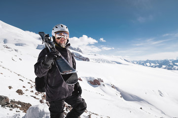 Fototapeta na wymiar Portrait bearded male skier aged against background of snow-capped Caucasus mountains. An adult man wearing ski googles mask and helmet skis on his shoulder looks mountains. Ski resort concept