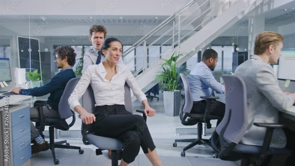 Poster cheerful employee pushes his beautiful female colleague on a chair between rows of desks with divers