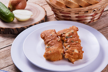 Deep-fried battered cod fillets, in a white plate next to onions, cucumber and bread on a wooden background. Close-up