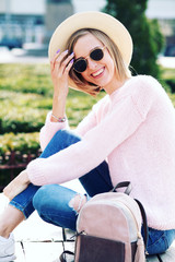 Image of traveling smiling woman walking in spring city. Hipster outfit. Oversize pink sweater, straw hat, round sunglasses and stylish accessories.