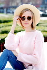 Image of traveling smiling woman walking in spring city. Hipster outfit. Oversize pink sweater, straw hat, round sunglasses and stylish accessories.