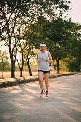 Young woman runner wears white sport vest, white cap, black short pant runs on the road to exercise in the evening while listens to music through earphone. Female runner runs on the city road.