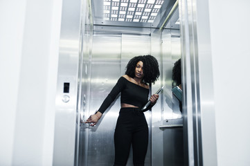 Fashionable young beautiful african american business woman with afro hairstyle wear in elegant black, stand at elevator with phone and laptop at hands.