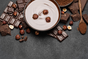 glass bowl of chocolate cream or melted chocolate, pieces of chocolate and hazelnuts on dark concrete background