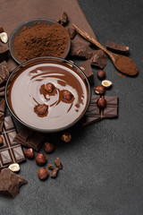 glass bowl of chocolate cream or melted chocolate, pieces of chocolate and hazelnuts on dark concrete background