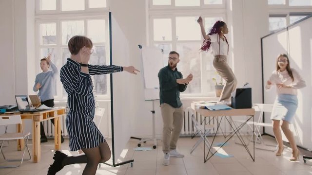 Young happy female office worker dancing together with multiethnic colleagues at fun corporate party slow motion.
