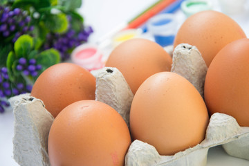 Easter brown chicken eggs in carton container, with plastic flowers and blurred paints, colorful palette on the background, paint equipment on white background