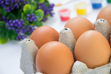 Easter brown chicken eggs in carton container, with plastic flowers and blurred paints, colorful palette on the background, paint equipment on white background