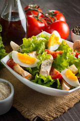 Fresh Caesar salad with delicious chicken breast, ruccola, spinach, cabbage, arugula, egg, parmesan and cherry tomato on wooden background. Oil, salt and pepper. Healthy and diet food concept.