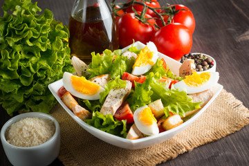 Fresh Caesar salad with delicious chicken breast, ruccola, spinach, cabbage, arugula, egg, parmesan and cherry tomato on wooden background. Oil, salt and pepper. Healthy and diet food concept.