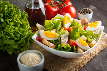 Fresh Caesar salad with delicious chicken breast, ruccola, spinach, cabbage, arugula, egg, parmesan and cherry tomato on wooden background. Oil, salt and pepper. Healthy and diet food concept.