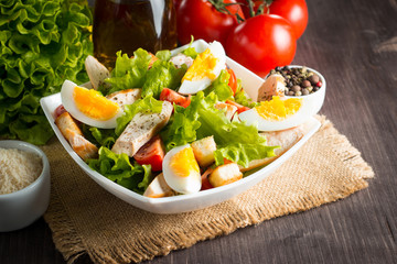 Fresh Caesar salad with delicious chicken breast, ruccola, spinach, cabbage, arugula, egg, parmesan and cherry tomato on wooden background. Oil, salt and pepper. Healthy and diet food concept.
