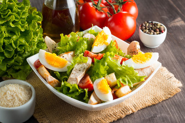 Fresh Caesar salad with delicious chicken breast, ruccola, spinach, cabbage, arugula, egg, parmesan and cherry tomato on wooden background. Oil, salt and pepper. Healthy and diet food concept.