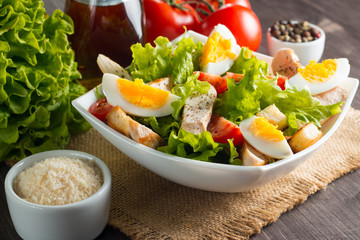 Fresh Caesar salad with delicious chicken breast, ruccola, spinach, cabbage, arugula, egg, parmesan and cherry tomato on wooden background. Oil, salt and pepper. Healthy and diet food concept.