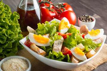 Fresh Caesar salad with delicious chicken breast, ruccola, spinach, cabbage, arugula, egg, parmesan and cherry tomato on wooden background. Oil, salt and pepper. Healthy and diet food concept.