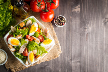 Fresh Caesar salad with delicious chicken breast, ruccola, spinach, cabbage, arugula, egg, parmesan and cherry tomato on wooden background. Oil, salt and pepper. Healthy and diet food concept.