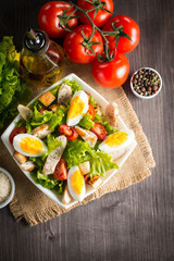 Fresh Caesar salad with delicious chicken breast, ruccola, spinach, cabbage, arugula, egg, parmesan and cherry tomato on wooden background. Oil, salt and pepper. Healthy and diet food concept.