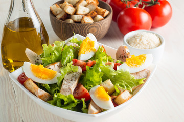 Fresh Caesar salad with delicious chicken breast, ruccola, spinach, cabbage, arugula, egg, parmesan and cherry tomato on wooden background. Oil, salt and pepper. Healthy and diet food concept.