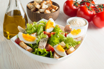 Fresh Caesar salad with delicious chicken breast, ruccola, spinach, cabbage, arugula, egg, parmesan and cherry tomato on wooden background. Oil, salt and pepper. Healthy and diet food concept.