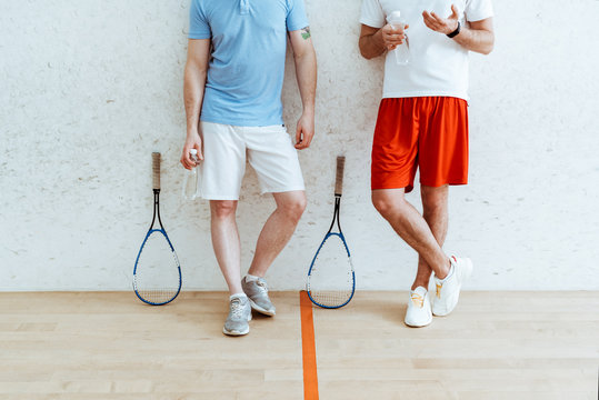 Cropped view of two squash players in shorts standing with crossed legs in four-walled court