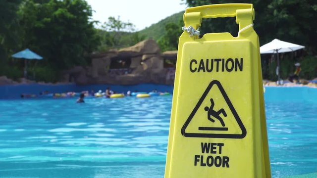 Yellow wet floor caution sign on swimming pool background in amusement water park. Caution wet floor sign in aqua park on swimming poolside background.