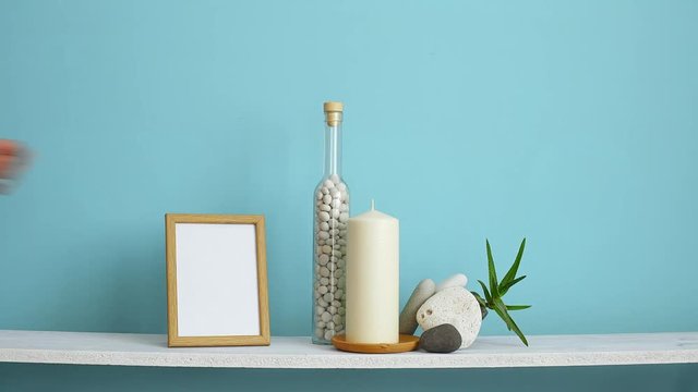 Modern room decoration with frame mockup. White shelf against pastel turquoise wall with Candle and rocks in bottle. Hand putting down empty picture frame.
