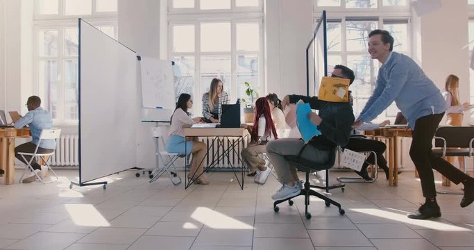 Dolly shot of crazy fun businessman riding office chair along office hall, celebrating and throwing paper in the air.