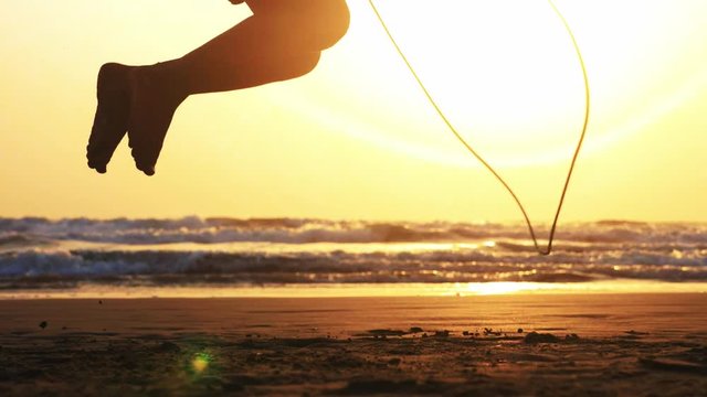 Silhouette of little girl's legs jumping on the rope on the sea sand beach at beautiful sunset, crop feet only, side view. Slow motion.