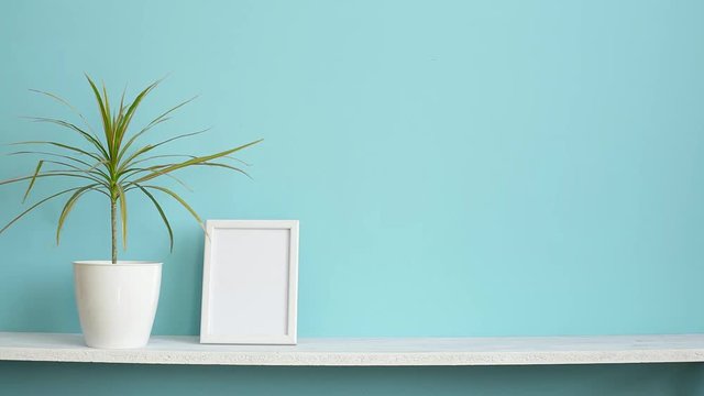 Modern room decoration with Picture frame mockup. White shelf against pastel turquoise wall and hand putting down potted dracaena plant.