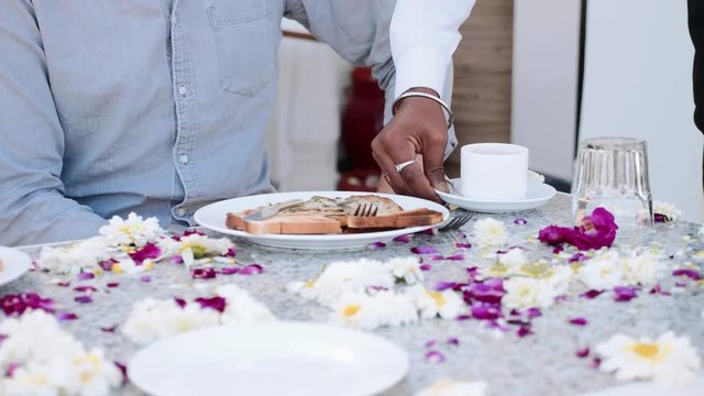 Waiter brings breakfast coffee to a couple sitting at the table covered with flowers petals in hotel. Table close-up. Slow motion.