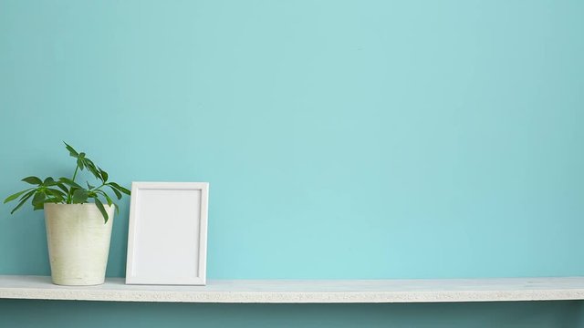 Modern room decoration with Picture frame mockup. White shelf against pastel turquoise wall and hand putting down potted schefflera plant.