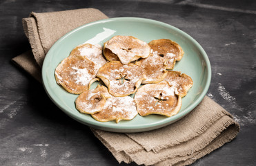 Apple chips sprinkled with powdered sugar and cinnamon on green plate on burlap on dark background.